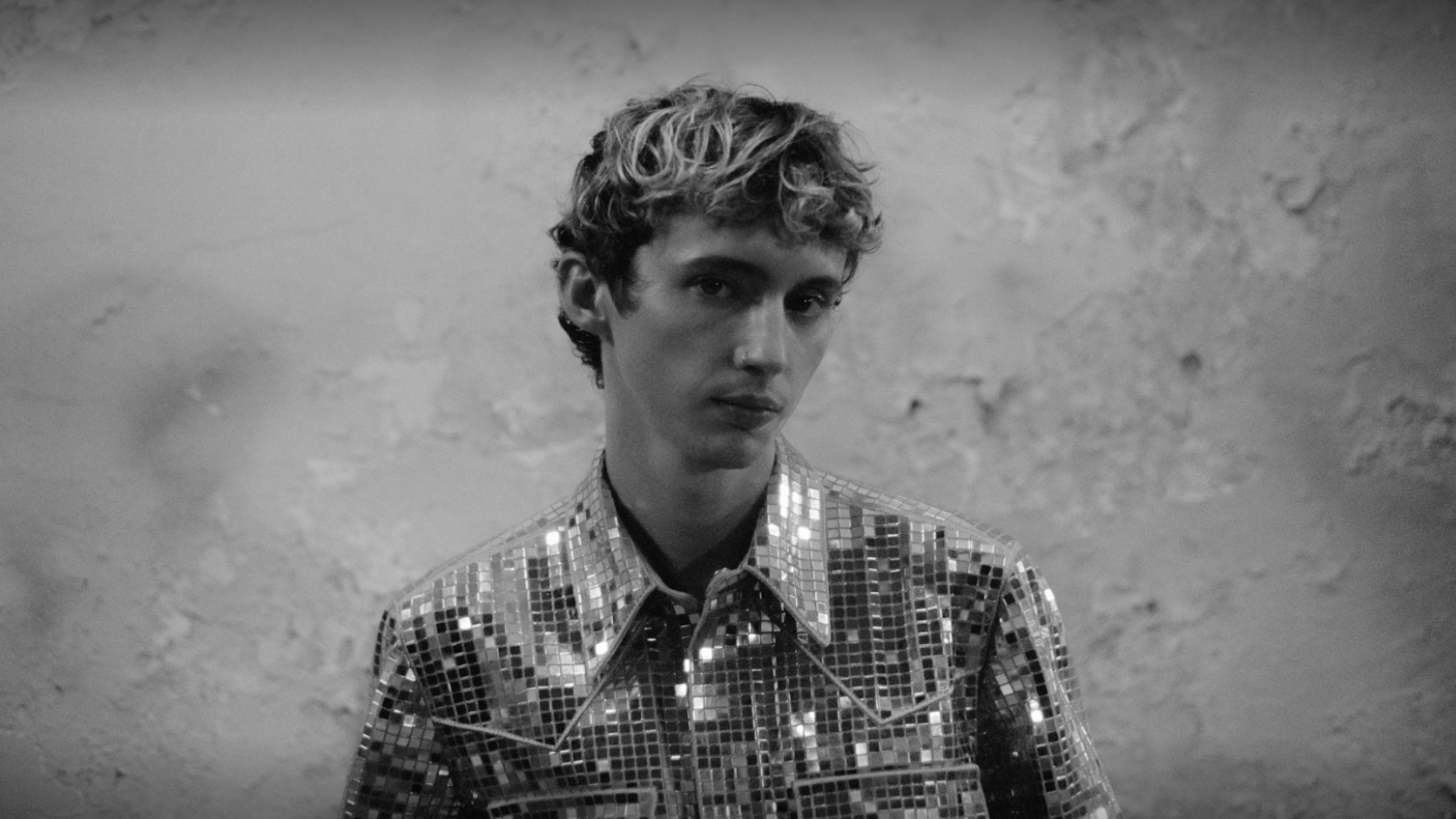 Troye Sivan in Schwarz-Weiß mit spiegelndem Hemd vor einer rauen Wand.
