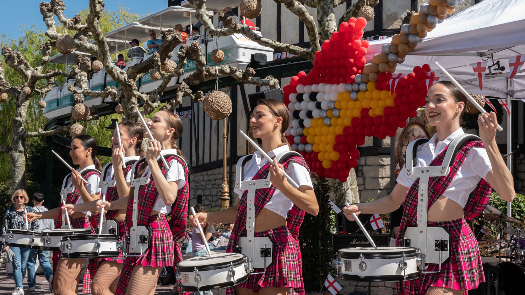 Mehrere Musikerinnen in rot karierten Outfits spielen mit Trommeln im englischen Themenbereich des Europa-Park. Im Hintergrund sind englische Flaggen, eine große farbige Ballondekoration und Teile der Parkkulisse zu sehen – passend zum Irischen und Englis