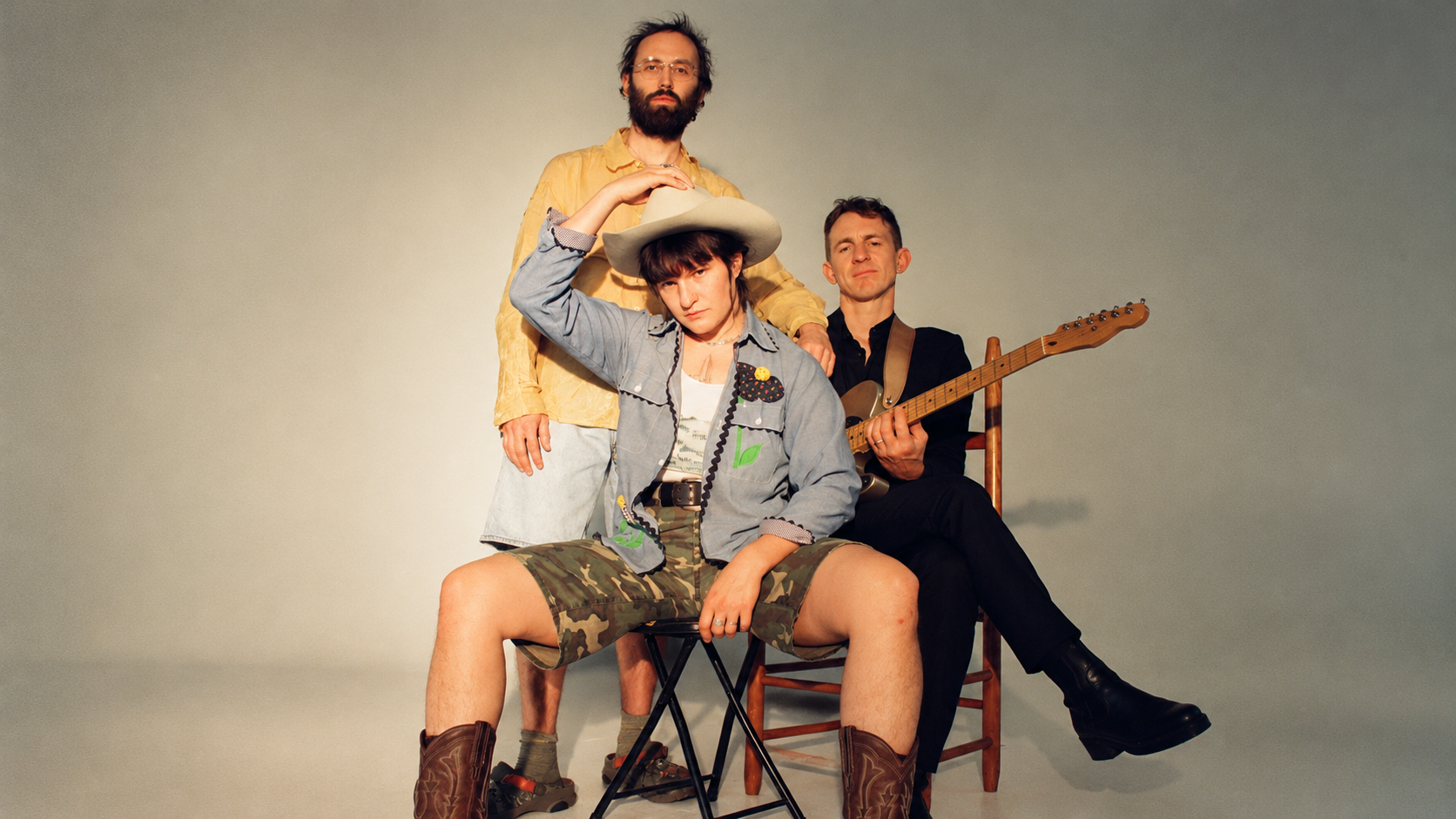 Drei Mitglieder einer Indie-Rockband posieren vor einer hellen Wand; eine Person sitzt mit Cowboyhut im Vordergrund, während zwei weitere Bandmitglieder dahinter stehen beziehungsweise mit einer Gitarre auf einem Stuhl sitzen.