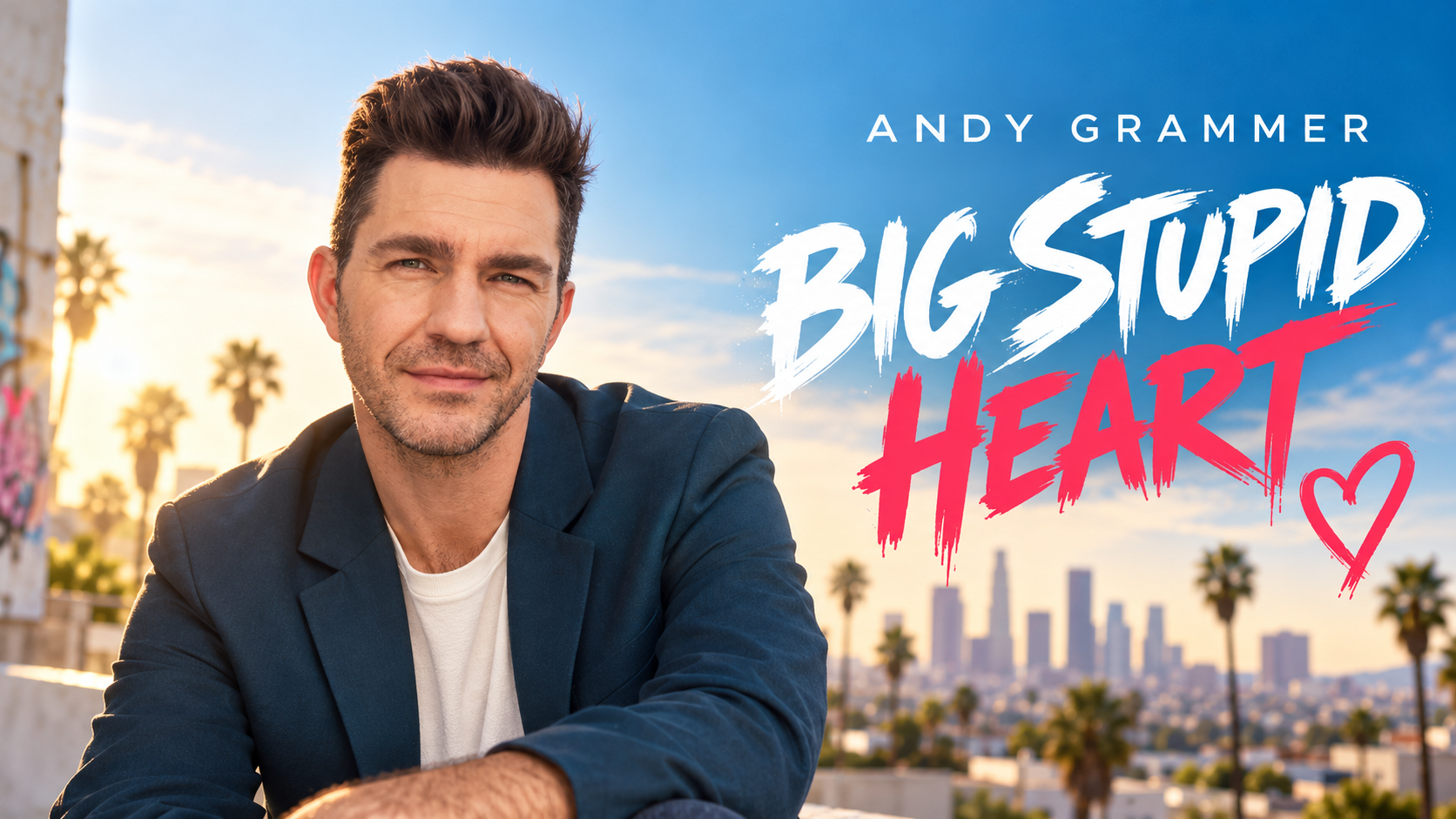 Andy Grammer sitzt in einem dunkelblauen Sakko vor einer sonnigen Stadtkulisse mit Palmen und Skyline; rechts ist der Schriftzug „Andy Grammer – Big Stupid Heart“ zu sehen.