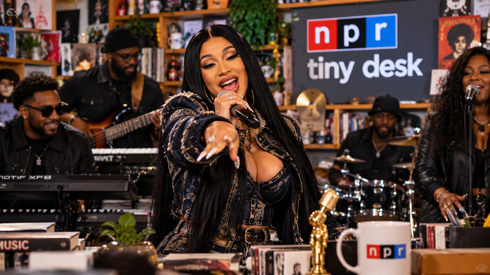 Redaktionelle Illustration von Cardi B bei einem intimen Tiny-Desk-Auftritt mit Mikrofon, Liveband, Instrumenten und dichtem Publikum in einem kleinen Studio.