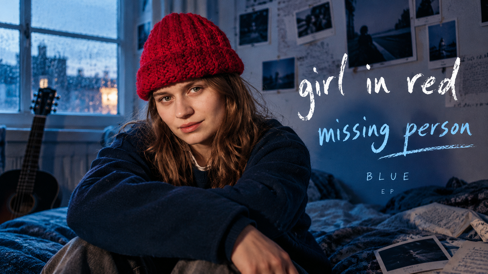 girl in red sitzt mit roter Strickmütze in einem blau beleuchteten Schlafzimmer zwischen Gitarre, Fotos und Notizen zur neuen EP „BLUE“.