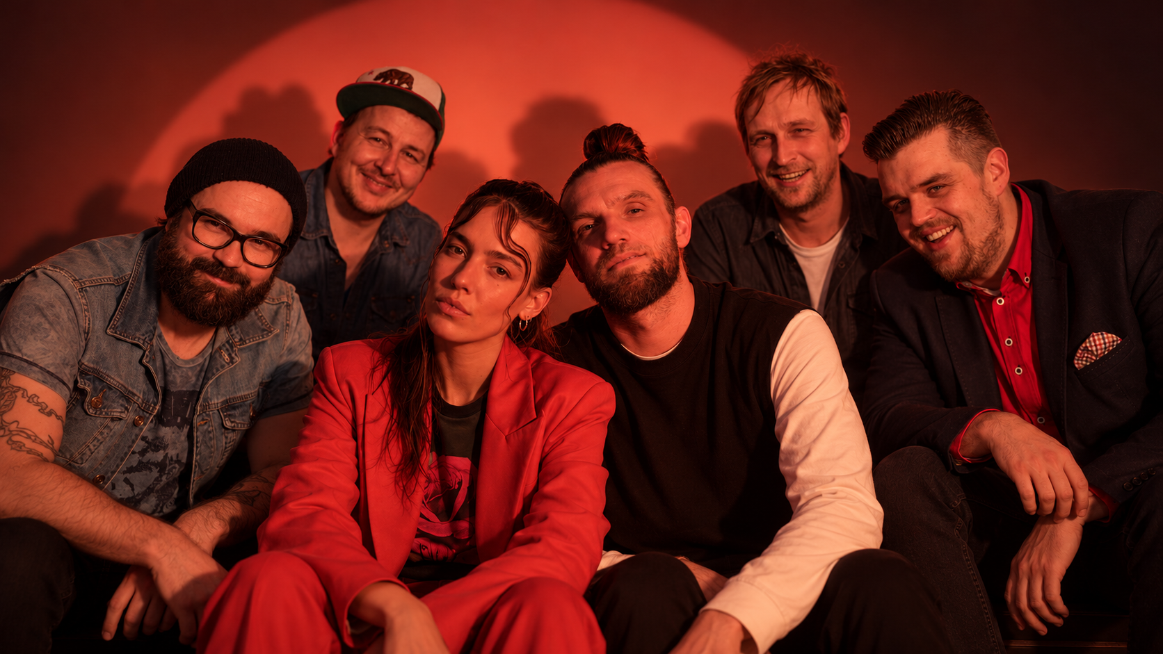 Frida Gold und Jupiter Jones posieren gemeinsam in einer warm rot beleuchteten Studioatmosphäre und bilden eine große gemeinsame Bandformation.
