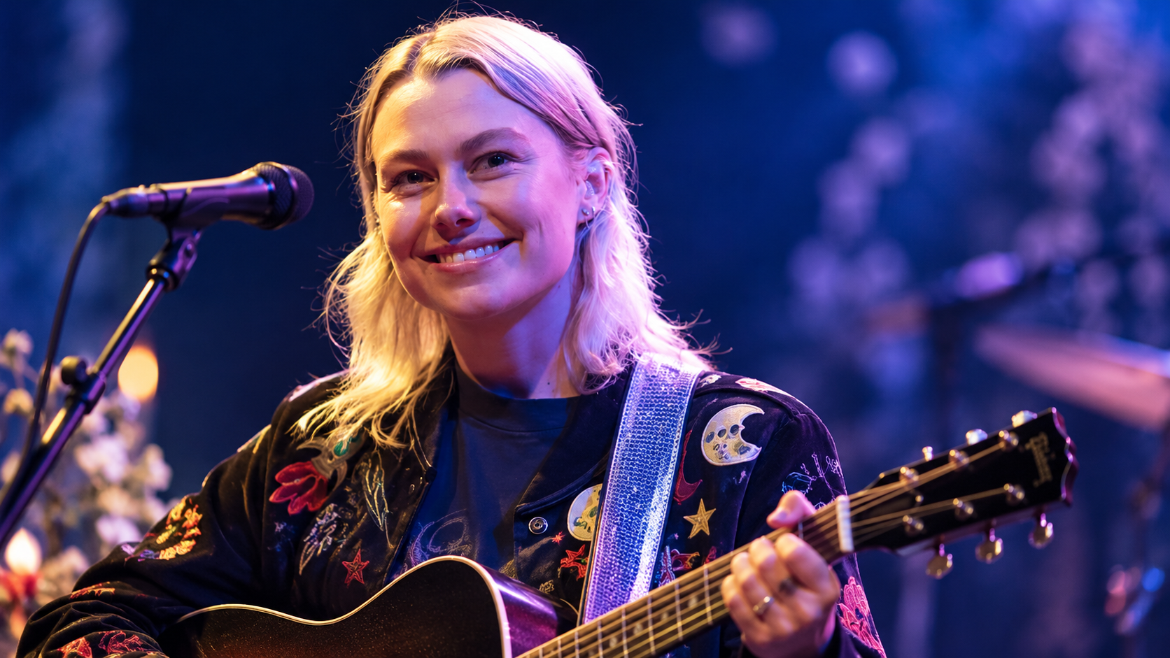Phoebe Bridgers steht mit einer Akustikgitarre auf einer Bühne und lächelt ins Publikum; blau-violettes Konzertlicht sorgt für eine atmosphärische Live-Stimmung.