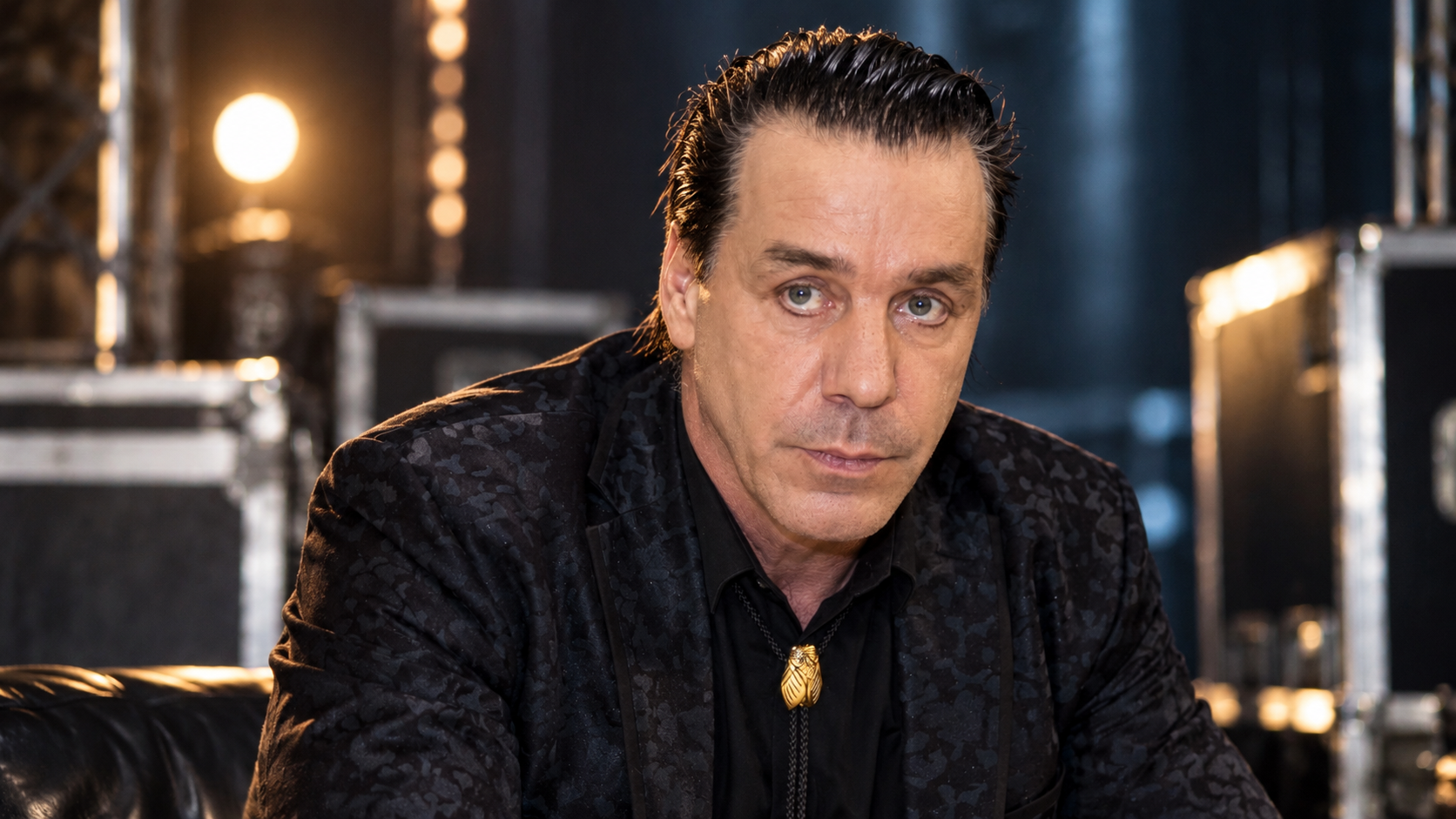 Till Lindemann sitzt in einem dunklen Backstage-Bereich zwischen Bühnen- und Tourcases; warmes Licht setzt sein Gesicht und sein schwarzes Outfit kontrastreich in Szene.