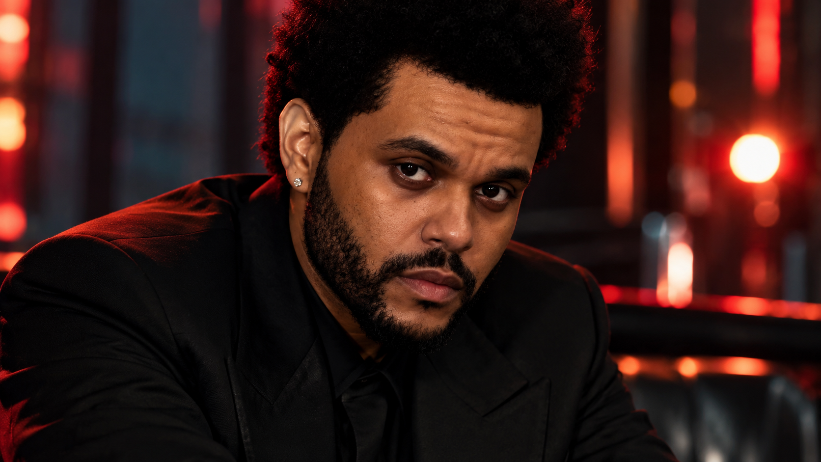 The Weeknd sitzt in schwarzem Anzug in einer dunkel beleuchteten Lounge; rotes und warmes Licht sorgt für eine intensive, elegante Atmosphäre.