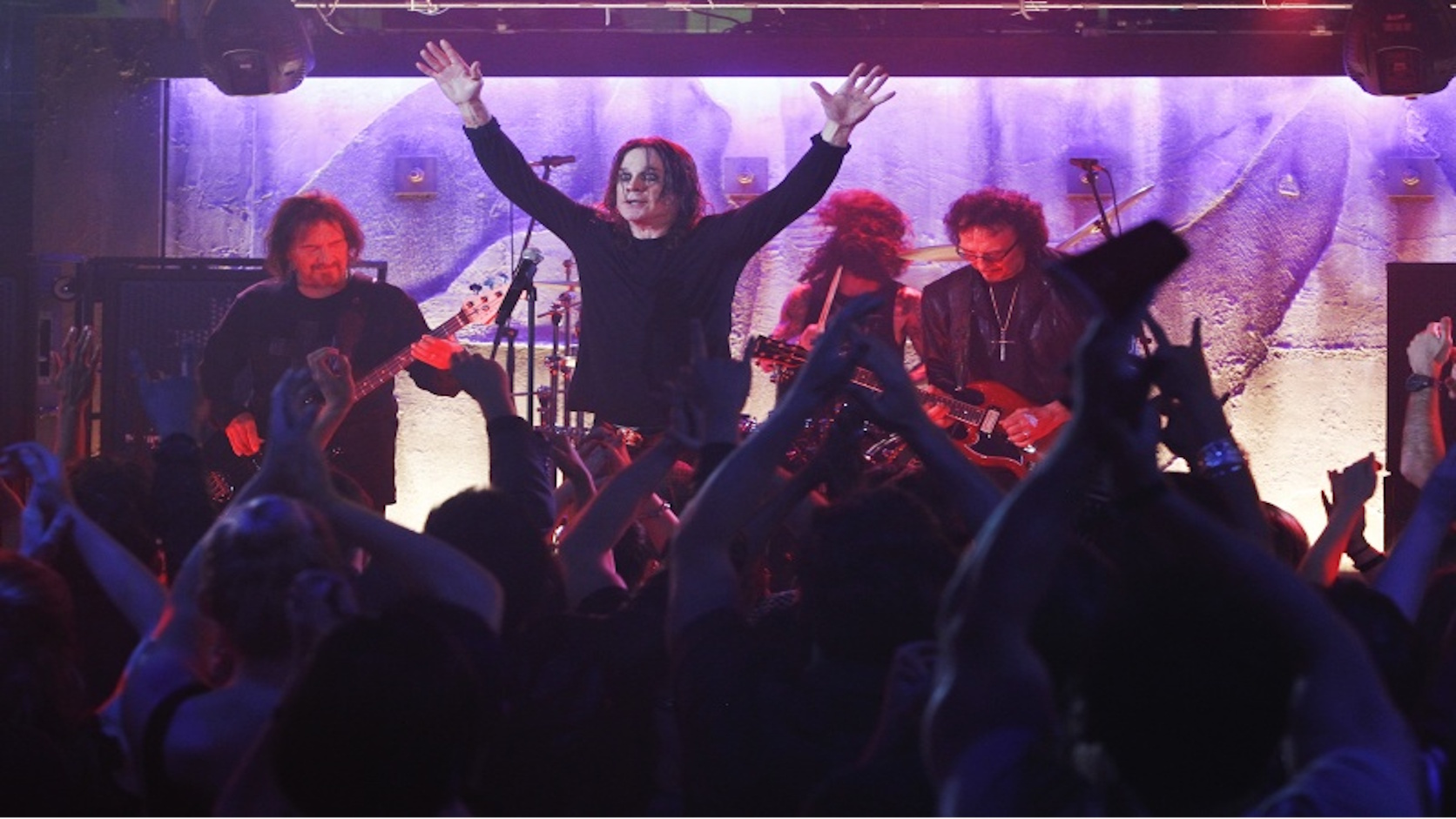 Ozzy Osbourne steht mit erhobenen Armen auf der Bühne, während Black Sabbath hinter ihm spielen und das Publikum im Vordergrund feiert.