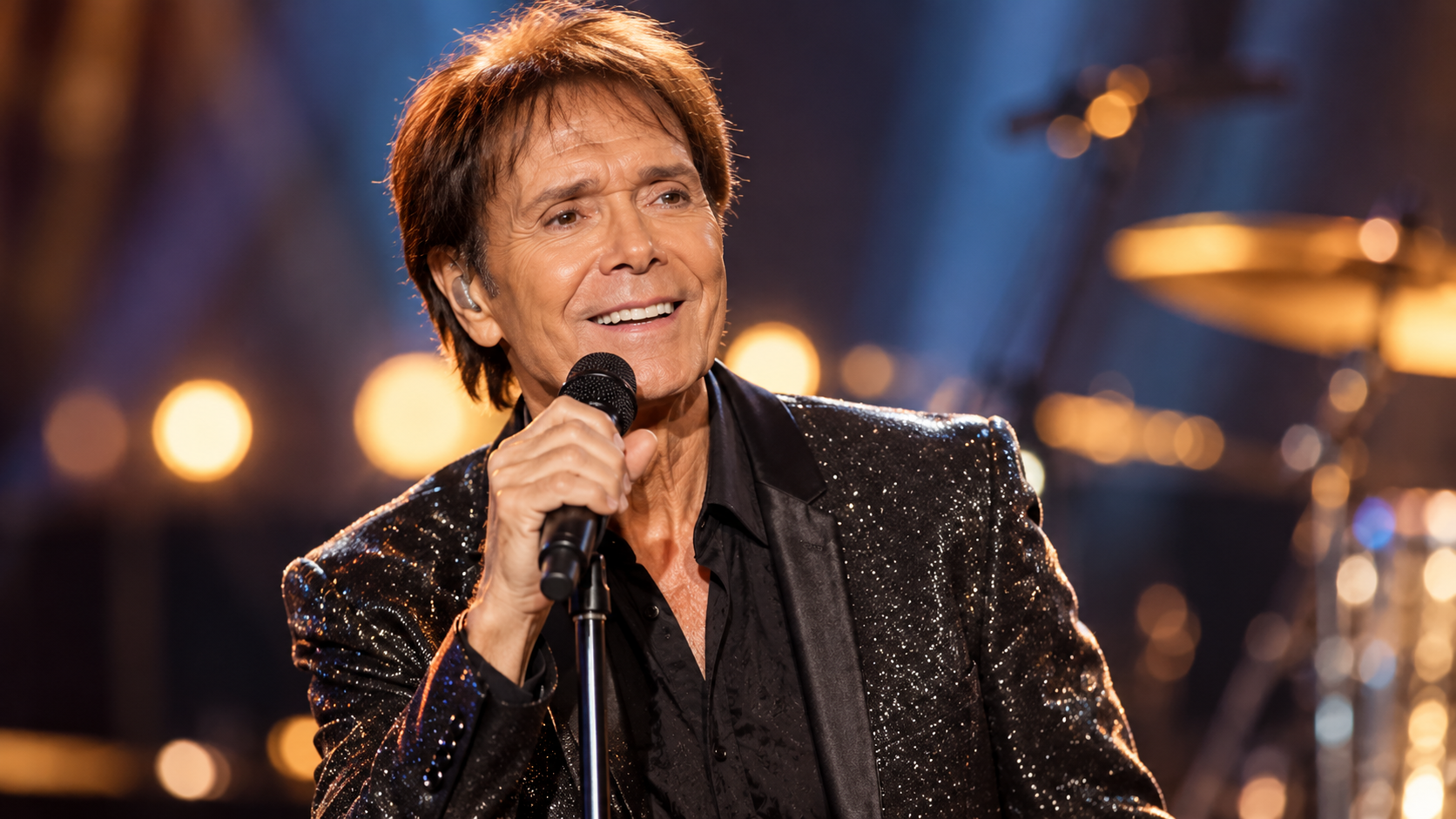 Cliff Richard steht mit Mikrofon auf einer Konzertbühne; warmes goldenes Licht und ein unscharfes Schlagzeug im Hintergrund sorgen für eine klassische Live-Atmosphäre.