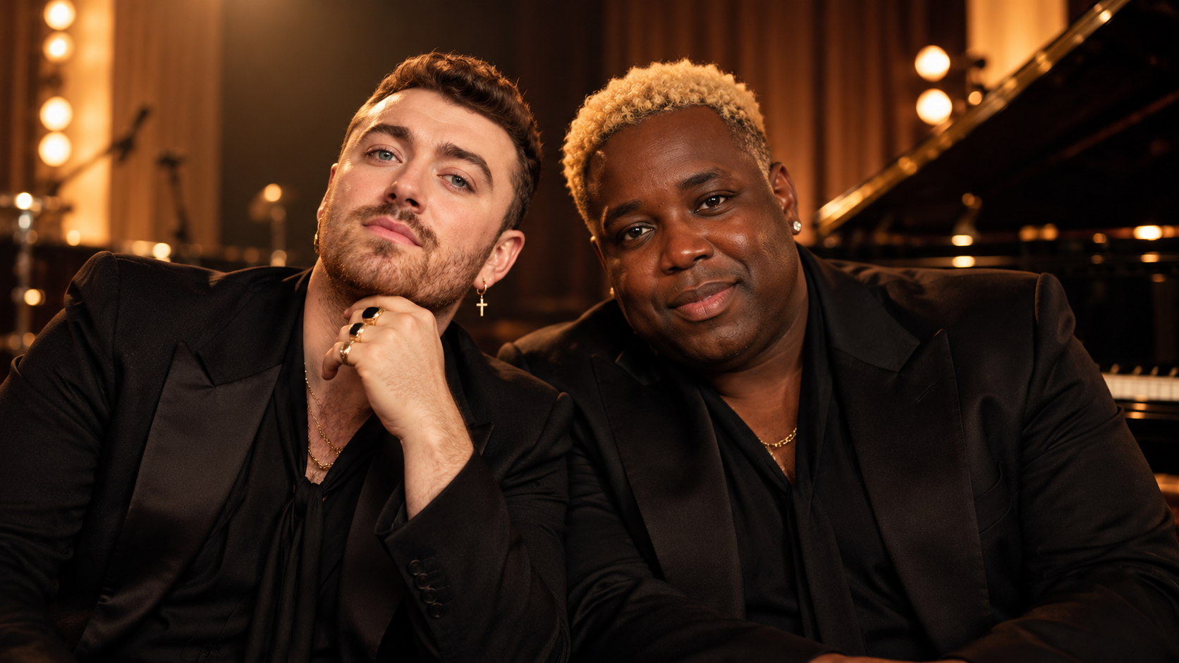 Sam Smith und Jacob Lusk sitzen gemeinsam in einem warm beleuchteten Studio neben einem Flügel; beide tragen elegante schwarze Outfits und blicken entspannt in die Kamera.